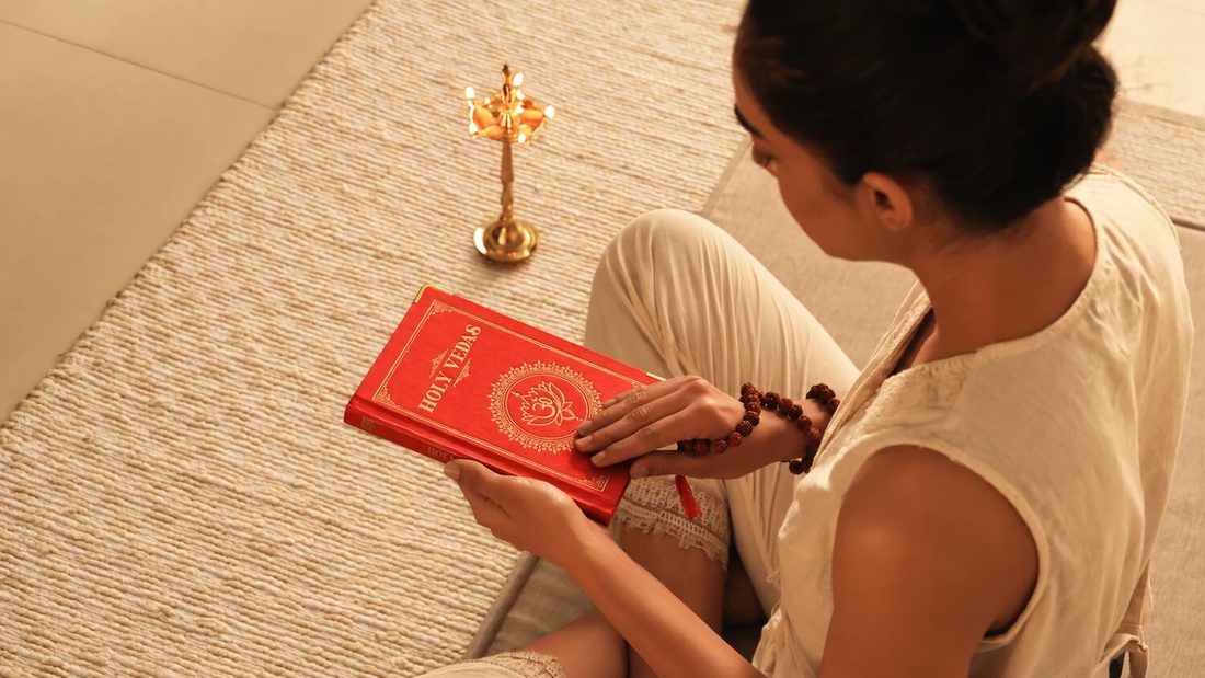 Ženska sedi na pleteni podlogi in bere rdečo knjigo Holy Vedas ob medeninasti ajurvedski oljni svetilki, za Art of Vedas